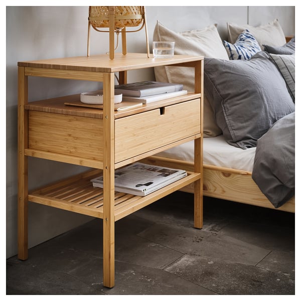 A bamboo NORDKISA bedside table with a drawer and open shelves. It holds books, a glass, and has a sleek, natural design.