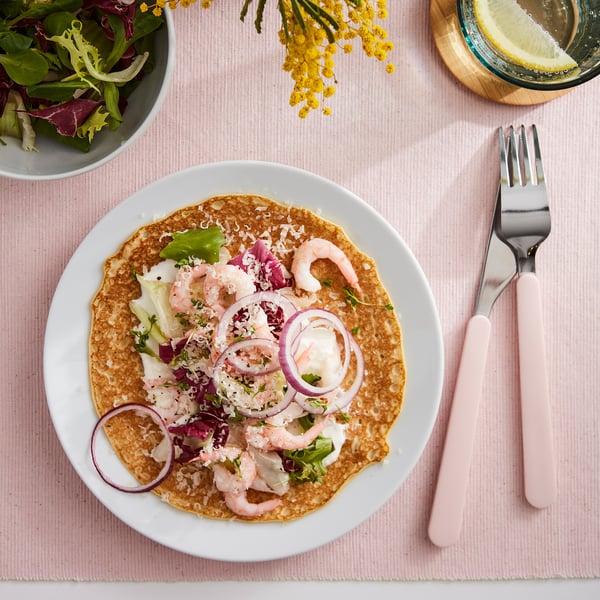 An overhead view of a savoury pancake topped with shrimp, red onions, and greens on a white plate next to pink cutlery and a bowl of salad.
