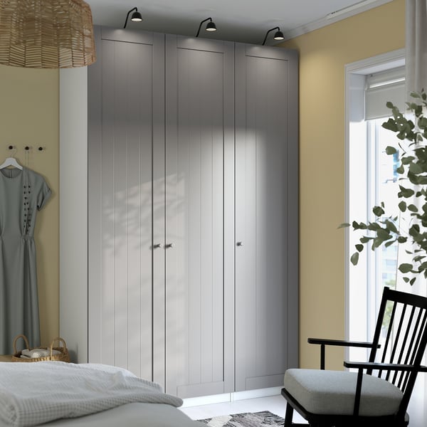 Modern bedroom with large grey PAX wardrobe, sleek design, tall and narrow, with lights and hangers, beside a cosy black chair.