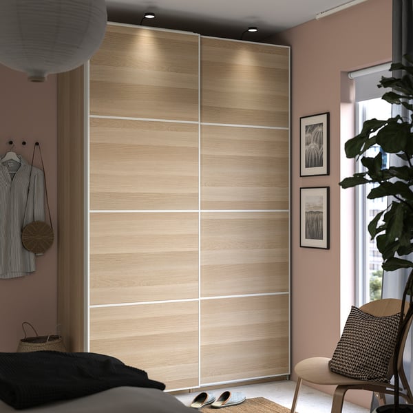 Modern bedroom with PAX wardrobe, sliding light-wood doors, hanging clothes, large plant, art prints, cosy chair.
