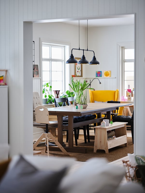 Bright dining room with a wooden table, yellow chairs, black bench, and hanging lamps.