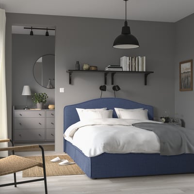 Modern grey bedroom with RAMNEFJÄLL blue bed, wooden dresser, wicker chair, hanging light, and shelf with books.