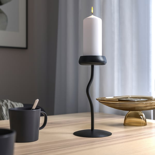 A lit white candle in a black, curvy candlestick on a wooden table, accompanied by black mugs and a gold plate.