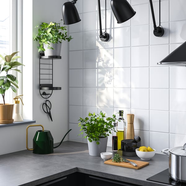 Modern kitchen with green plant stand, potted plants, white tiles, dark counter, steel stove, black lamps, lemons, herbs, tools.