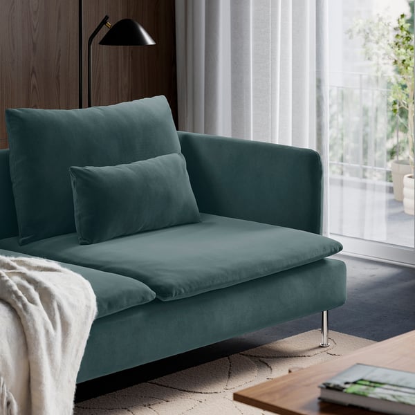 A teal sofa with two pillows and metal legs sits by a window with white curtains. A black floor lamp is nearby. A white throw rests on the sofa.