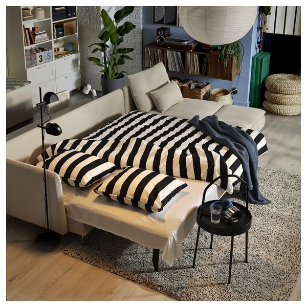 Modern living room with beige sofa-bed, black & white striped bedding, rug, side table, bookshelf, plants, and large hanging lamps.