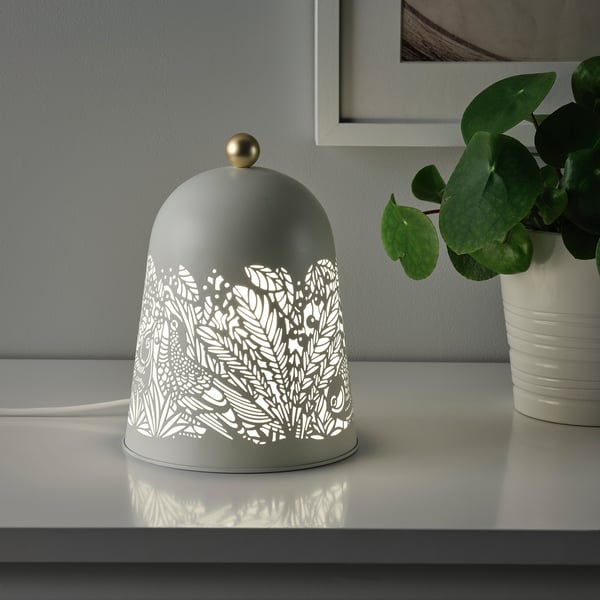 A decorative table lamp, SOLSKUR, with a nature-inspired design provides soft mood lighting, sitting on a white surface next to a potted plant.