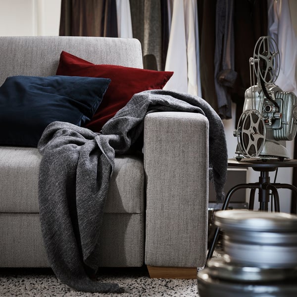 Modern grey SÖRVALLEN sofa with red and blue pillows, draped with a grey blanket, next to a film projector on a stand.