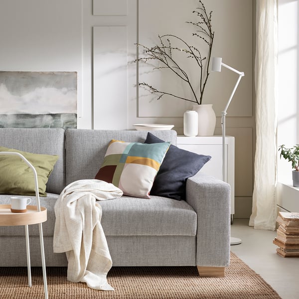 Modern grey 3-seat sofa with colourful pillows, white side table, lamp, and plants in bright room.