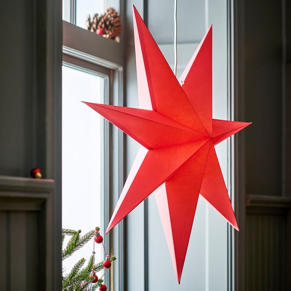 Large red paper star pendant light, ctrl hangs prominently near window, surrounded by small christmas tree with red ornaments.