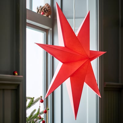 Large red paper star pendant light, ctrl hangs prominently near window, surrounded by small christmas tree with red ornaments.