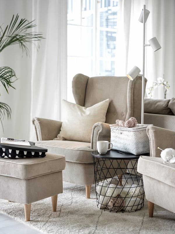 Modern living room with beige STRANDMON wing chair, striped ottoman, metal side table, and plants.