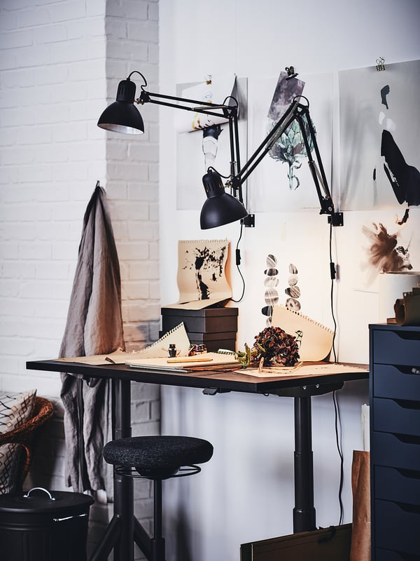 Contemporary home office with TERTIAL black adjustable desk lamp, white bricks, art posters, grey fabric, and wooden stool.