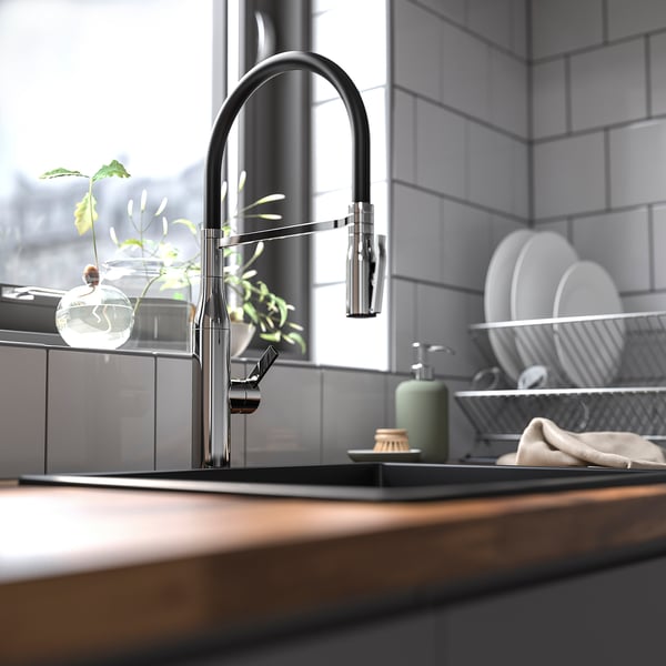 Modern kitchen with TOLLSJÖN chrome tap, sleek and tall, near a window. Wooden counter, grey tiles, plants, and white dishes in a rack beside it.
