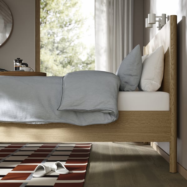 Bedroom with wooden bed frame, light blue bedding, slippers on striped rug near window.