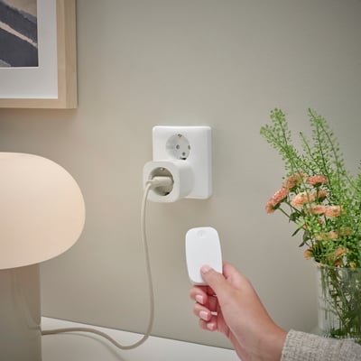 Hand holding remote controls smart plug; connected lamp, flowers, wall socket.