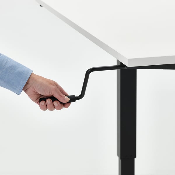 Image shows a person adjusting an adjustable standing desk with a crank handle, switching between sitting and standing heights.