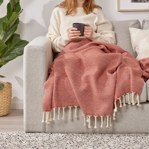 A person relaxes on a sofa, wrapped in a soft, blush-colored wool throw, sipping from a mug. The throw fringe details, enhancing comfort and warmth.