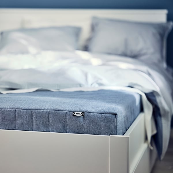 Close-up of IKEAs VADSÖ blue-gray mattress on white bed, showing fabric cover and foam top.