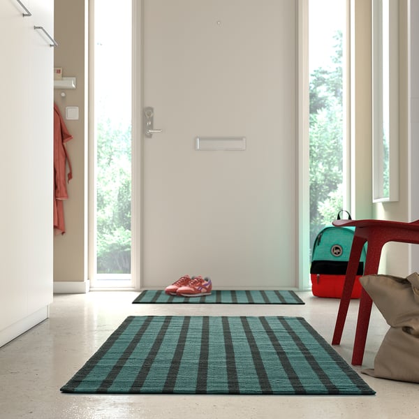 Modern entryway with large blue-green plaid rug, smaller matching mat, red kids chair, and green backpack near white doors.