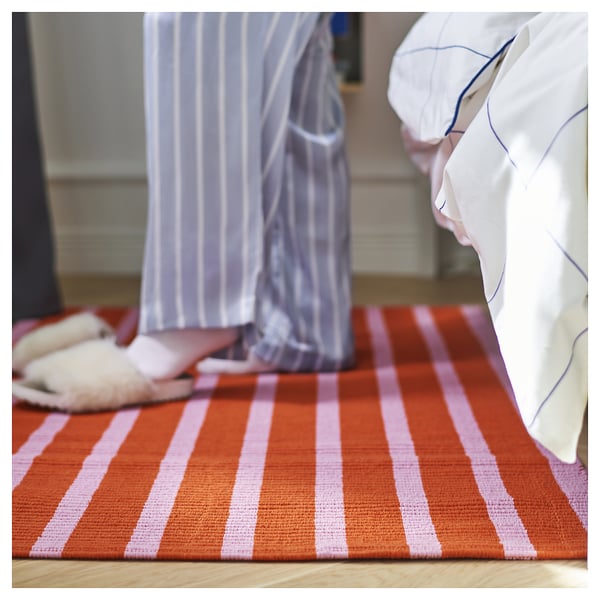 A VÄGSKYLT rug, featuring pink and orange stripes, made of cotton. It is rectangular, flat, and designed for easy vacuuming.