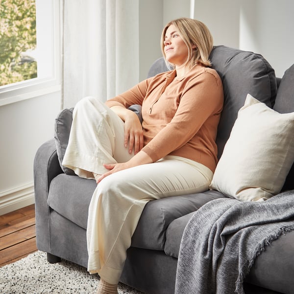 Relaxed person sits on large grey VINLIDEN sofa, beige blanket on legs, soft cushions, natural light from window, calm setting.