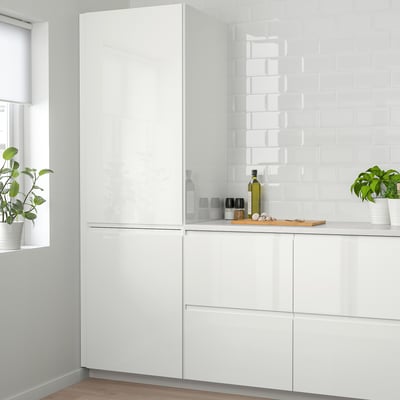 A modern white kitchen with glossy VOXTORP cabinets and backsplash. Potted plants and olive oil bottle are placed on the countertop.