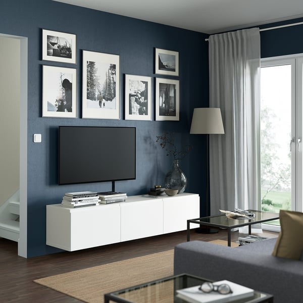 Modern living room with dark blue walls, white tv bench, flat-screen tv, five black & white photos, tall lamp, glass coffee table, grey sofa.