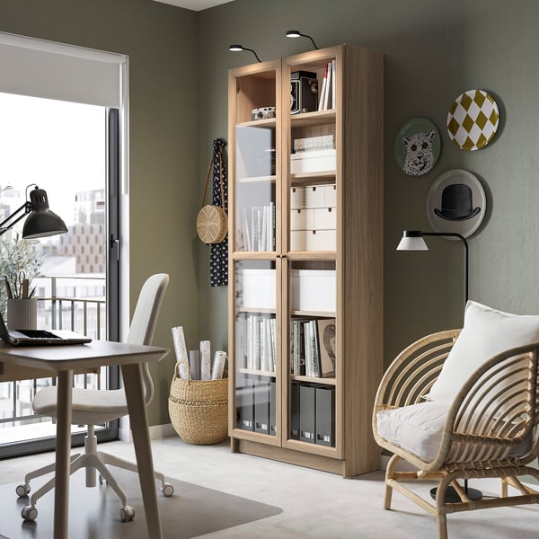 Light wooden BILLY bookcase with glass doors, near a white chair & small table, decorated with plates and vases.