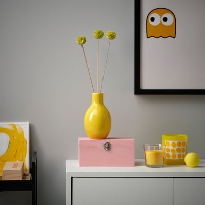 Bright yellow BLODBJÖRK vase with round shape holds three flowers. Placed on pink box near glassware and artwork, highlights cheerful decor.