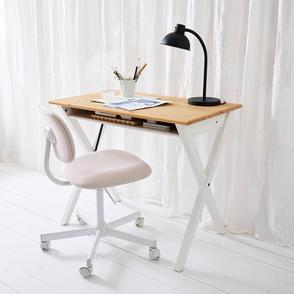 A blonde BOLLPOJKE desk with papers, lamp, and chair. White curtains brighten the room.