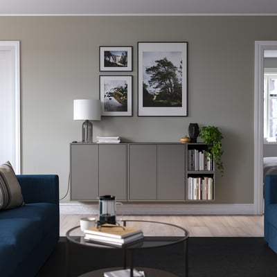 Modern living room with blue sofa, round table, and grey EKET storage. Wall decor includes framed photos and a plant.