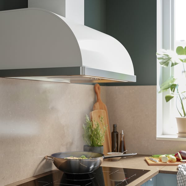 Modern kitchen with white range hood, stove, and wooden counter; greenery, cooking pans, and apples nearby.