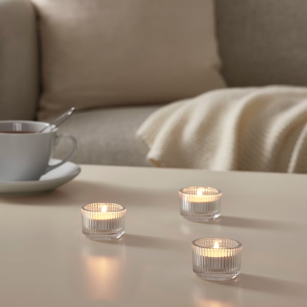 Three lit GLIMMA candles on white table, blue cup, spoon, beige blanket on couch, warm glow.