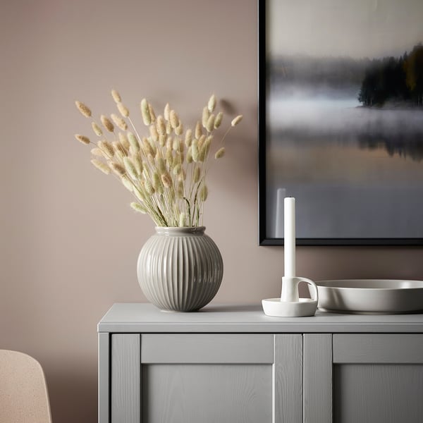 A minimalist, modern candlestick named GODTAGBAR, white in colour, made of stoneware, standing small and sleek beside a large vase on a grey surface.