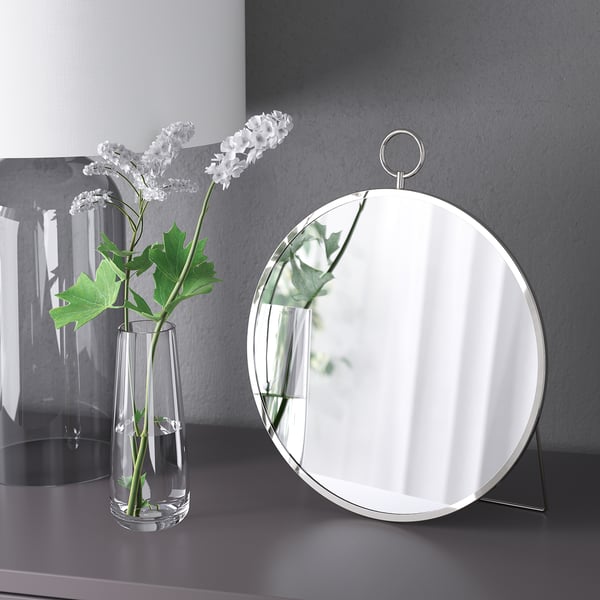 A round mirror on a stand next to a clear vase with white flowers. The mirror can hang or stand.