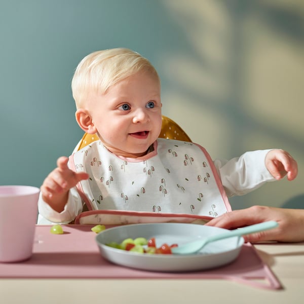 Baby wearing a floral bib sits in high chair, eating.