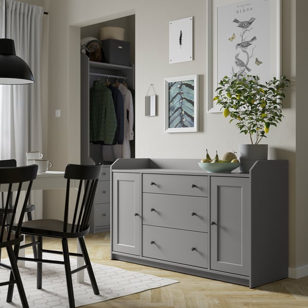Modern grey sideboard in living room, with drawers and doors, next to a closet with clothes. Decorated with art and a plant.