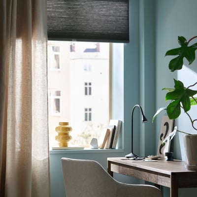 Home office with HOPPVALS light grey roller blind, wooden desk, lamp, vase, books, grey chair, and green plant.