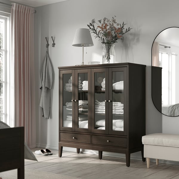 Modern brown wooden cabinet with glass doors, storing folded towels. Topped with a lamp and flowers, beside a mirror and coat hooks.