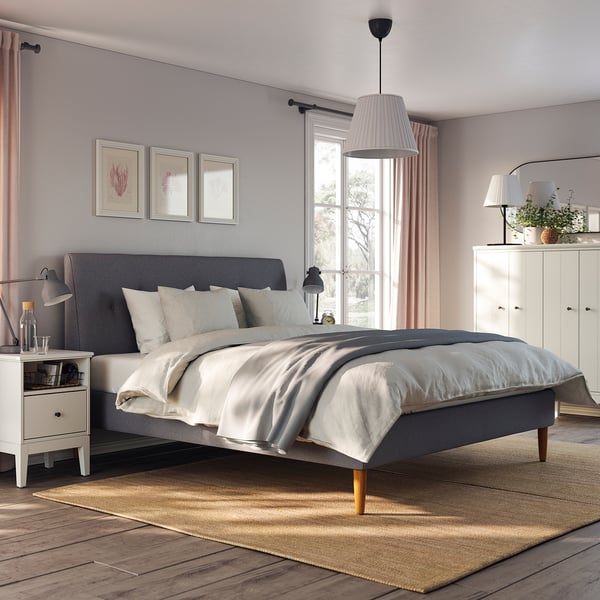 Modern bedroom with IDANÄS grey upholstered bed, white nightstands, large window, and natural rug, creating cosy and stylish ambiance.