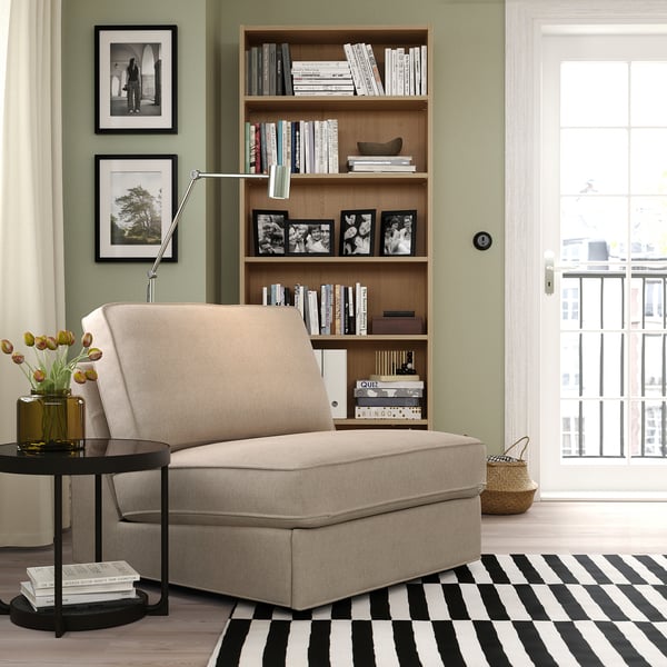 Modern living room with sage walls, a beige KIVIK sofa-bed, striped rug, and wooden bookshelf filled with books and black and white photos.