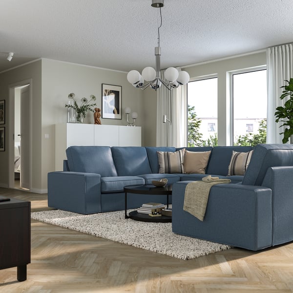 Modern living room with blue KIVIK sectional, chandelier, and coffee table. Decor includes rug, plants, art, and large windows.