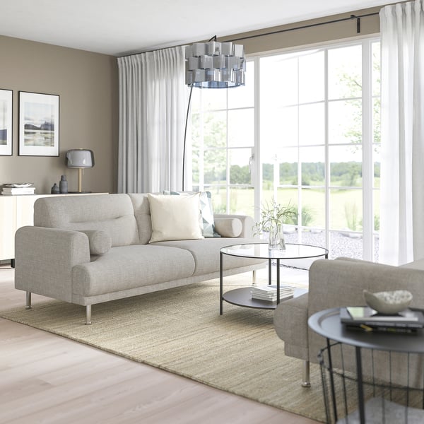 Modern living room with LÅNGARYD sofa, glass table, grey curtains, lamp, and books. Large windows flood it with natural light.