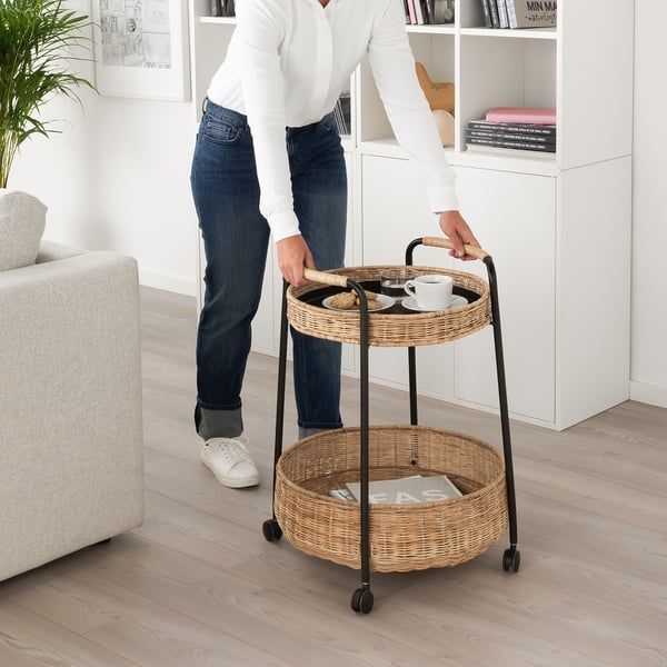 Person pushes a two-tiered rattan cart with a tray and basket. Ideal for serving and storing items at home.
