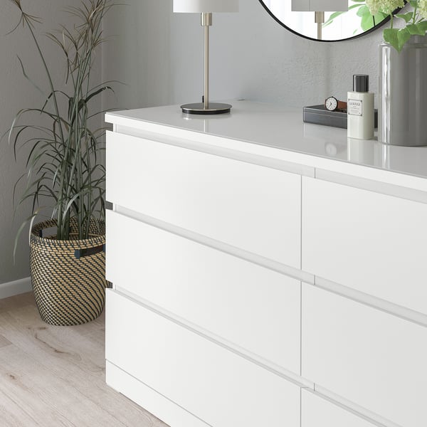 White chest of drawers with glass top, protecting surface. Small lamp, watch, and bottles on top, plant beside.