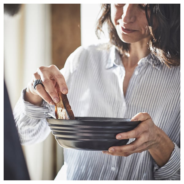 A person wearing a striped shirt holds a black, round bowl, dipping a piece of bread.