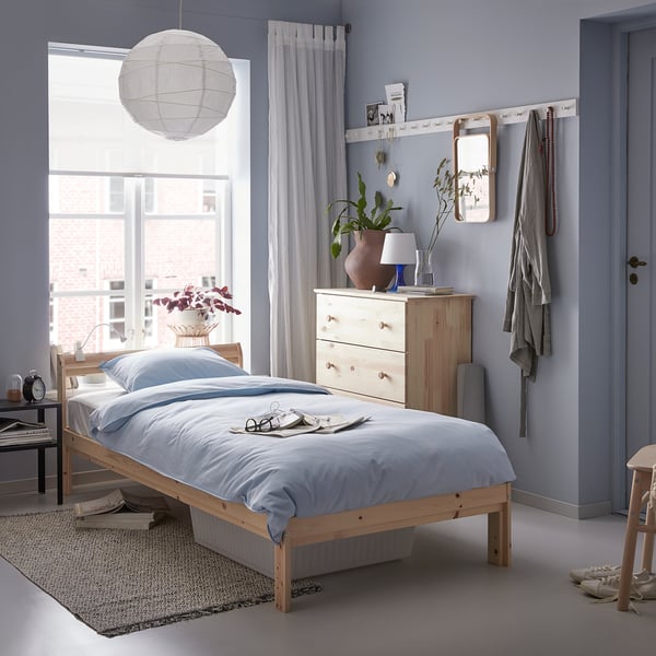 Minimalist bedroom with NEIDEN light wood bed, light blue linens. White pendant lamp, sheer curtains, wooden dresser, and plants.