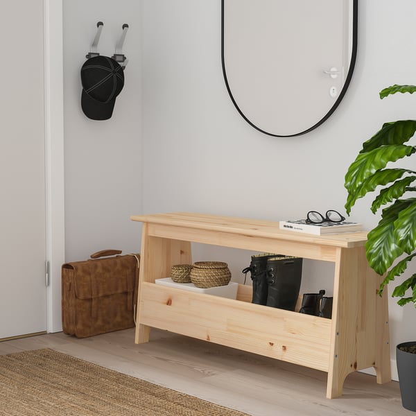 A brown bench with woven basket, black bag, mirror, green plant, and tan bag nearby.