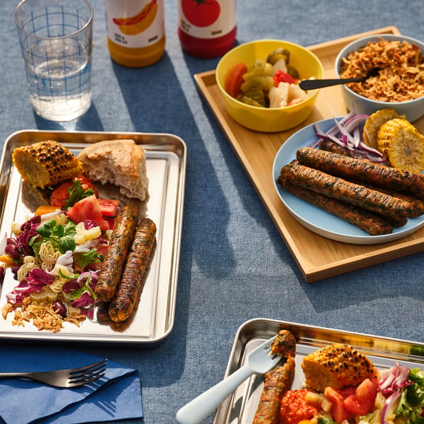 Four plates with grilled sausages, one plate with salad, bread, and tomatoes. Two bowls of mixed veggies and sauerkraut, all on a blue tablecloth.